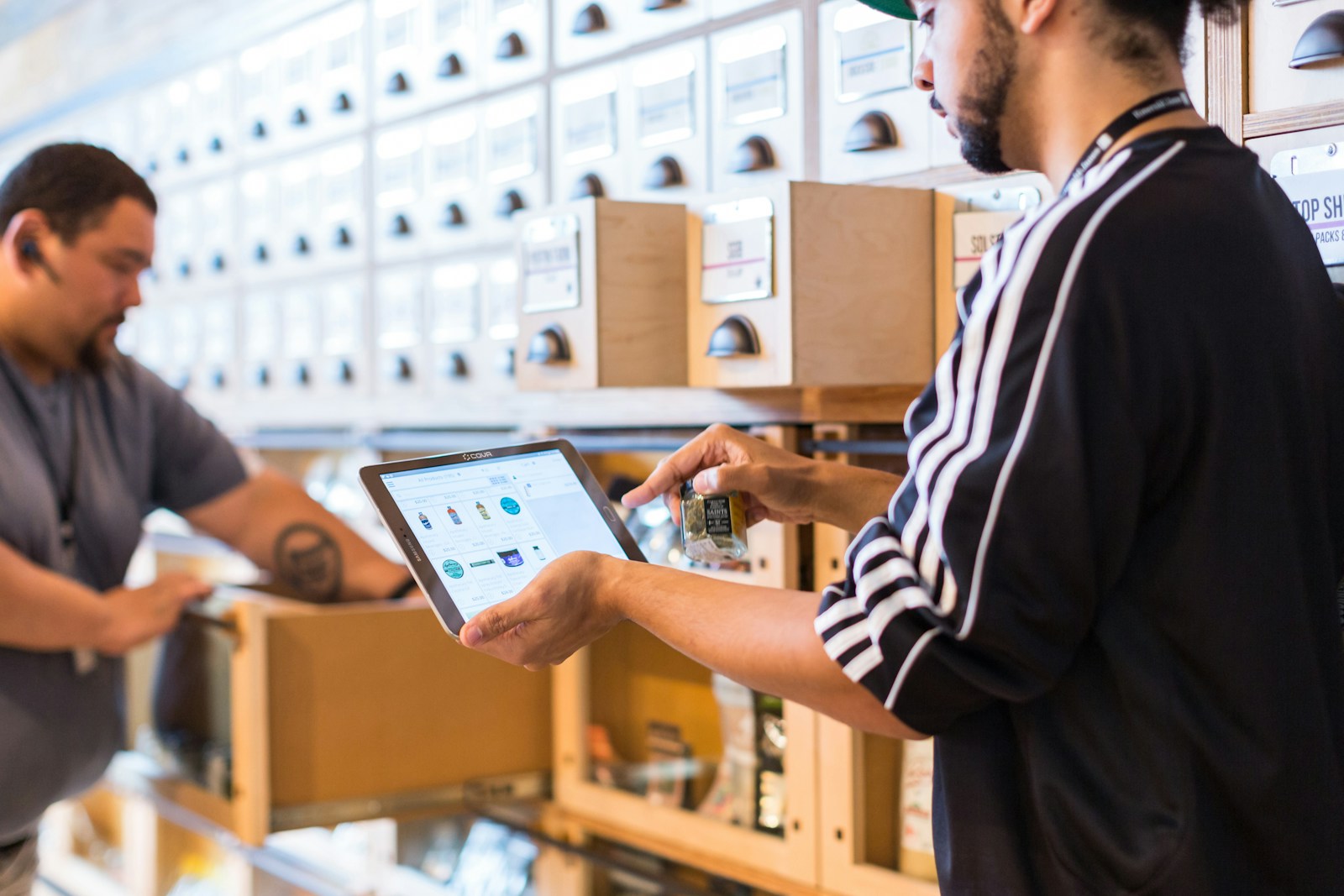 A dispensary employee uses a point-of-sale tablet while another worker manages inventory