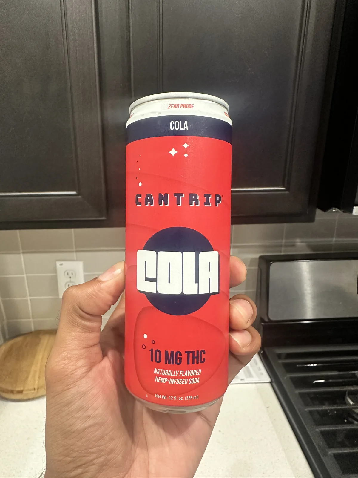 A hand holding a red Cantrip Cola can labeled Zero Proof, 10 MG THC, naturally flavored hemp-infused soda, in a home kitchen