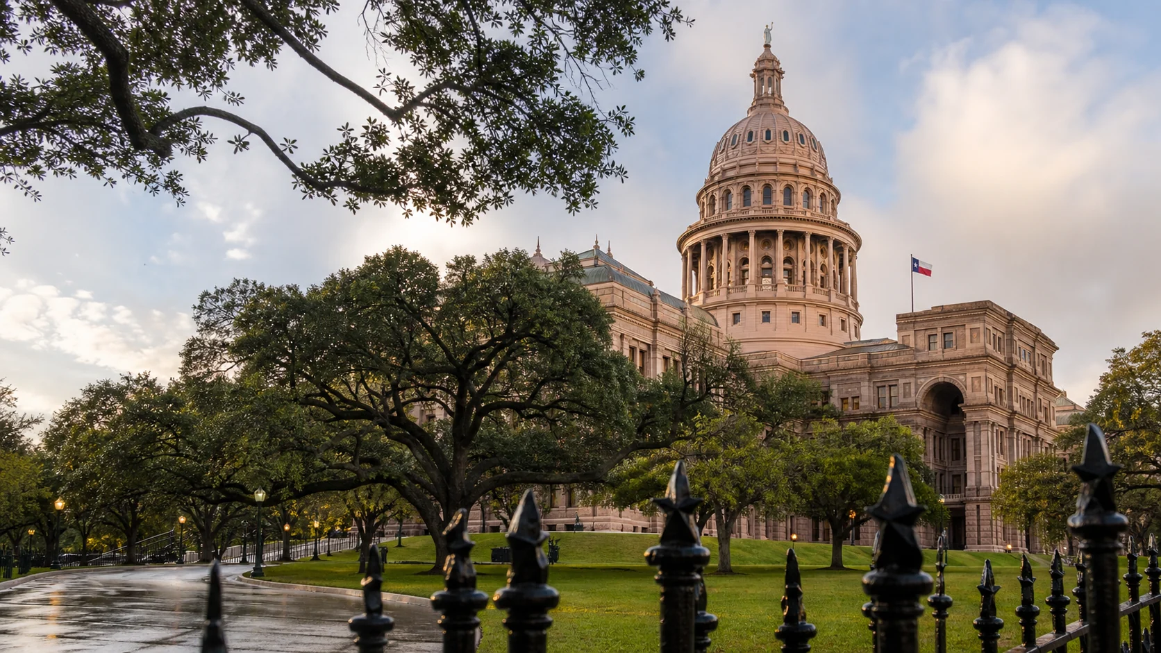Editorial view of the Texas State Capitol framed by live oak trees and a wrought-iron fence